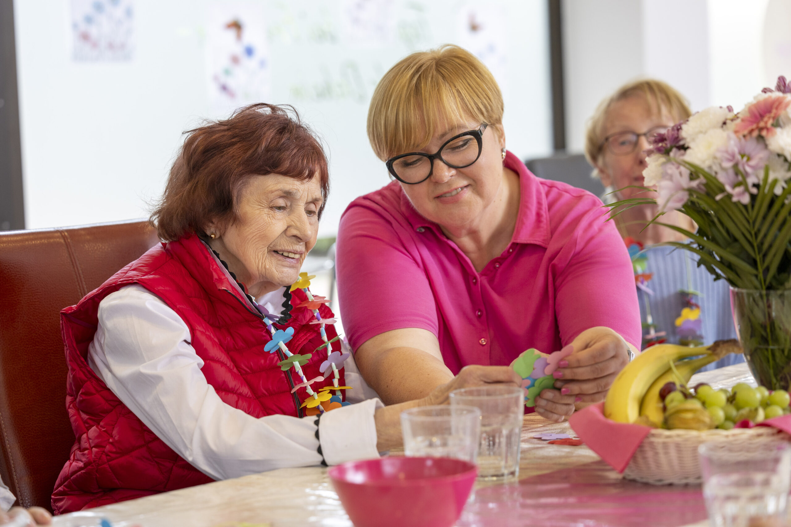 Mitarbeiterin und Seniorin sitzen am Tisch und sprechen miteinander; kleine Beschäftigung liegt bereit.