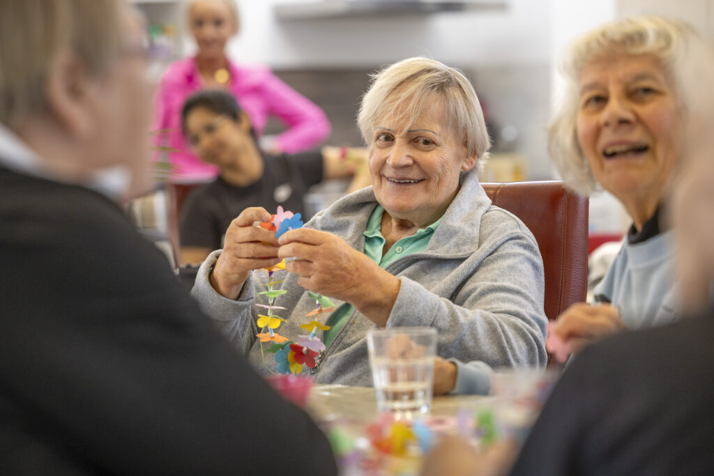 Gast sitzt am Tisch und fädelt bunte Elemente auf, Mitarbeitende begleiten aufmerksam in hellem Raum.