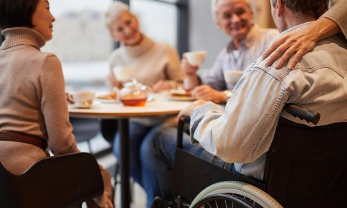 Gruppe älterer Menschen sitzt beim Kaffee am Tisch; Hand auf Schulter eines Rollstuhlfahrers – freundliche Gemeinschaft.